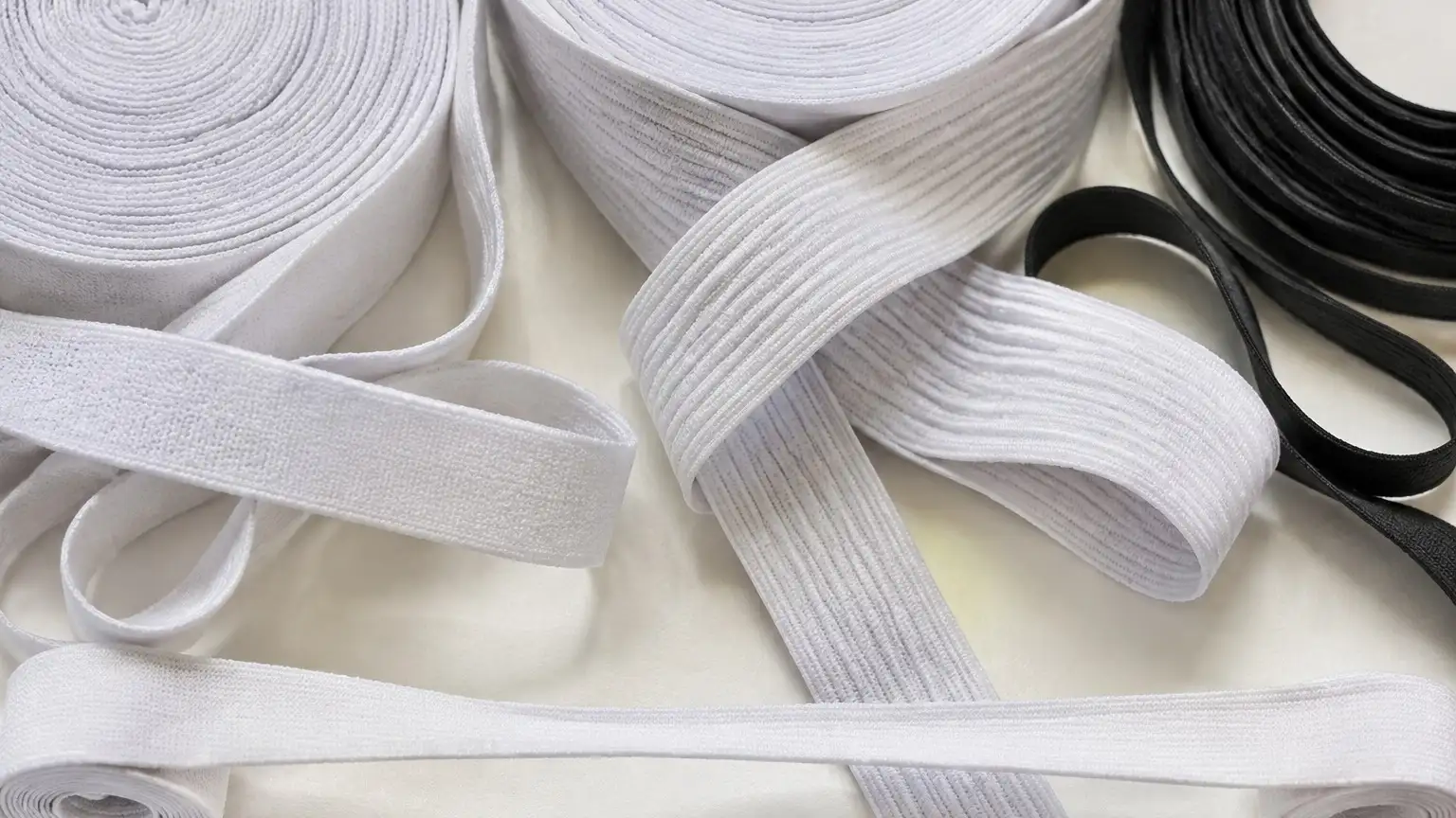 Rolls of white knitted and ribbed woven waistband elastic, partly unrolled, with a coil of narrow black elastic beside them.