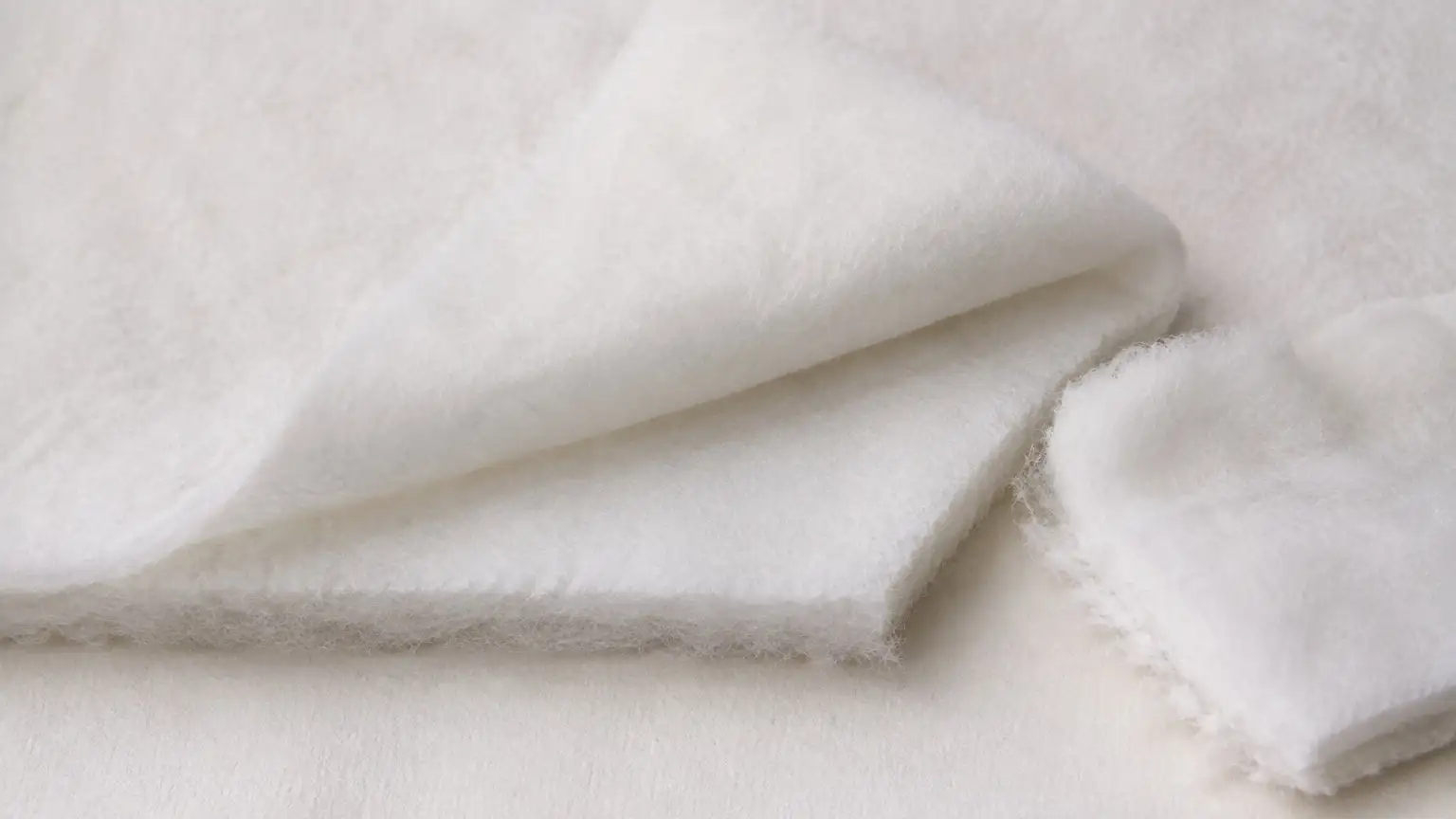 A thick soft sheet of white polyester wadding with one corner folded back to show its loft, and a torn piece showing the fibrous interior.